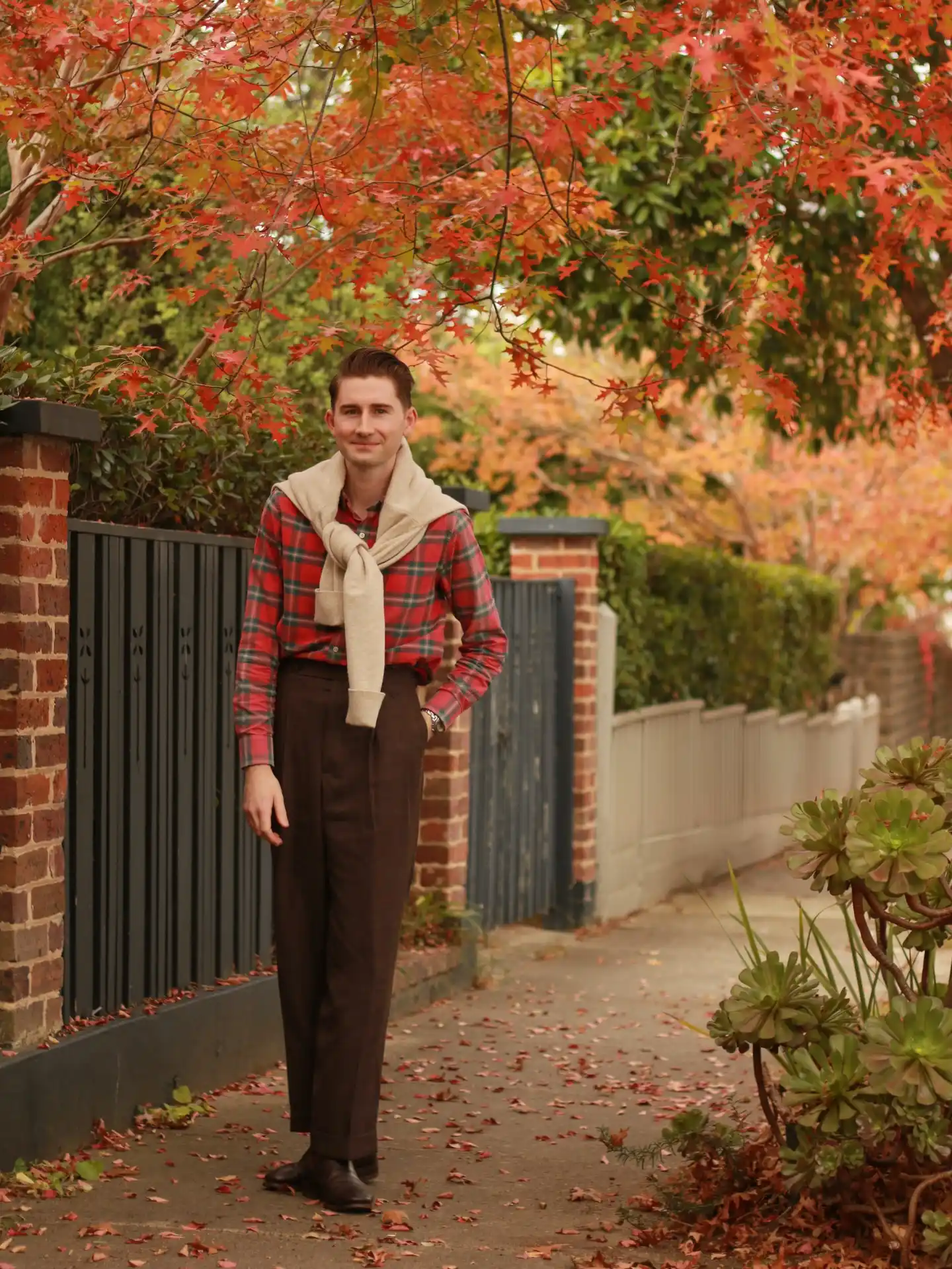 2. Plaid Shirt and Sweatershoulder Layering
This autumn ensemble focuses on a rich red and green plaid flannel shirt. Dark brown high waisted trousers establish a strong contrast at the waistline. A light beige sweater is draped over the shoulders, adding soft, contrasting texture without full bulk.Tying the sweater diagonally creates an immediate horizontal break across the chest. It disrupts the long line of a tall body. The high waisted pants clearly define the torso while the plaid draws the eye.