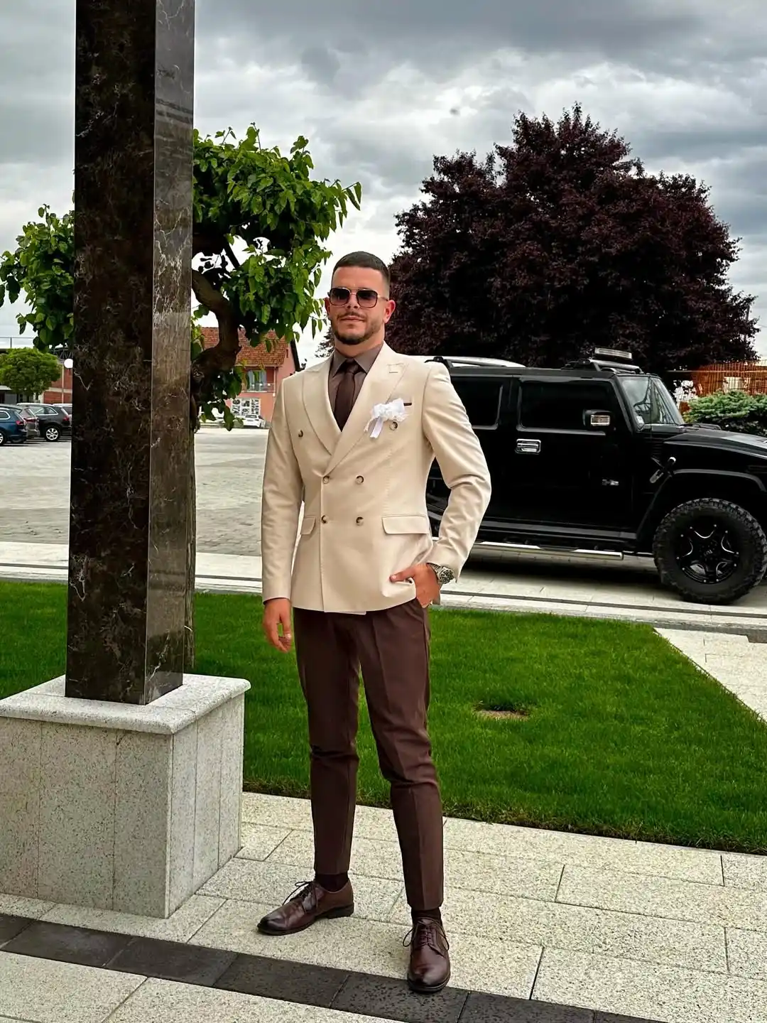 1. Cream Double Breasted Blazer With Brown Trousers
A cream double breasted blazer sets the tone here, paired with slim brown trousers that taper down to the ankle. Gold buttons add a subtle shine against the light fabric, while a white pocket square peeks out for contrast. Brown lace up shoes finish the look and tie the whole palette together.
Tall frames carry this two-tone combo well since the elongated blazer line pairs naturally with fitted trousers. Dark sunglasses and a slim brown tie complete the outfit, giving it a polished edge suited for daytime celebrations or garden ceremonies.