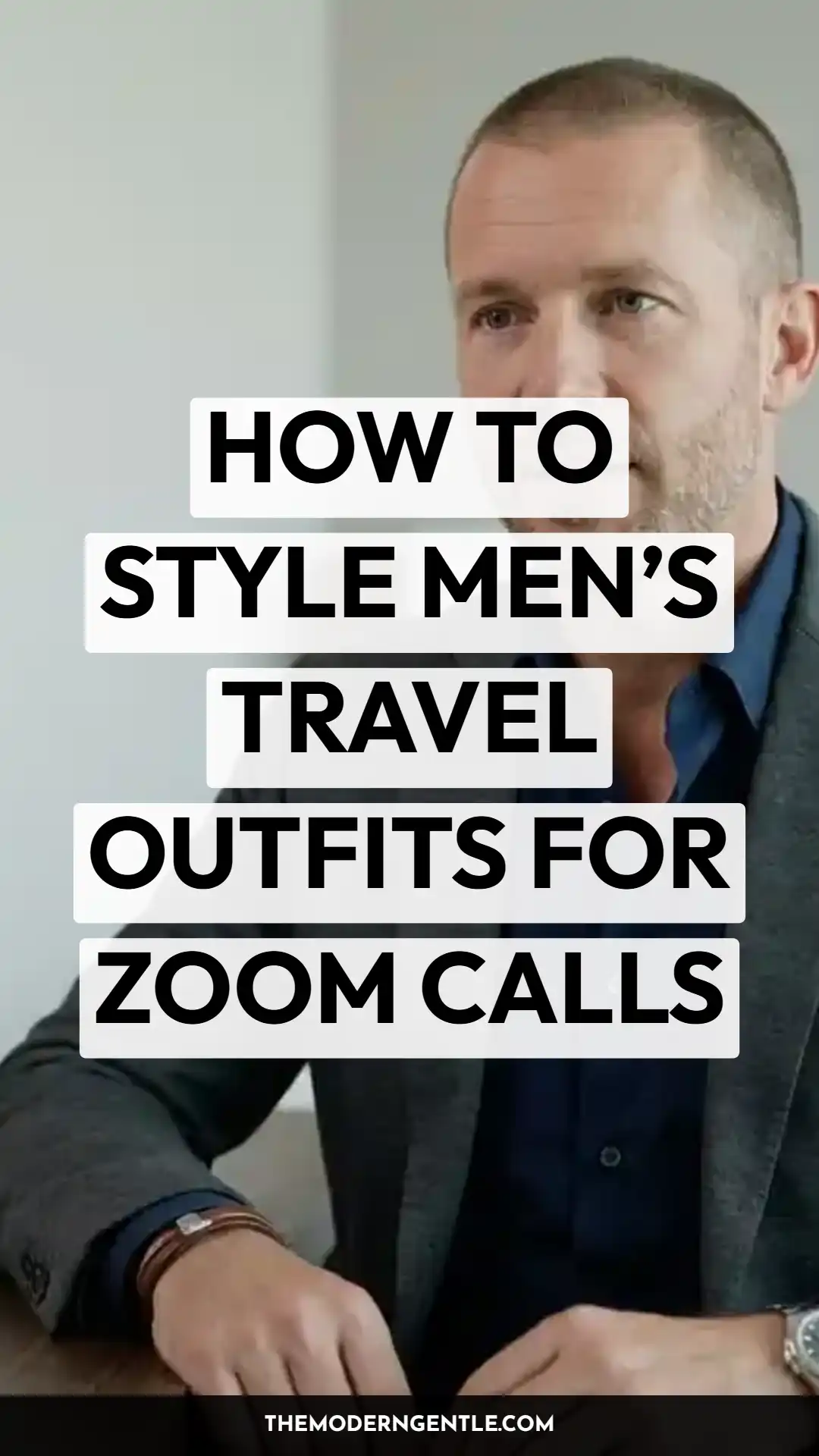 Close portrait of a man with short hair and light stubble wearing a heather grey blazer over a navy button-down shirt, a woven bracelet, and a watch, hands resting on a wooden table against a plain grey wall. Bold black text on white blocks reads "How to Style Men's Travel Outfits for Zoom Calls," illustrating business casual men's style for work travel and video call outfits.
