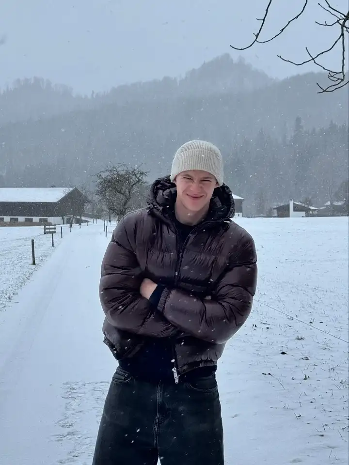 25 Stylish Winter Travel Outfits for Men & Ski Trips 6 Glossy Brown Puffer and a Cream Beanie in Falling Snow
