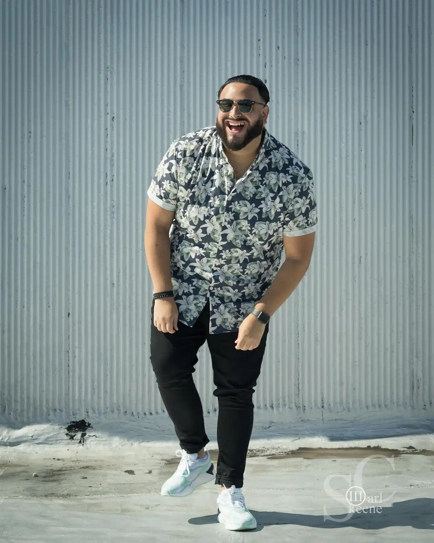 plus size men fashion11. Floral Print Button Up ShirtA man laughs mid stride in front of a corrugated metal wall, wearing a black and white floral button up shirt. Short sleeves and a relaxed collar give the piece an easy, breathable feel for warmer days. Beaded and leather bracelets stack on one wrist, adding a small personal touch.Black joggers taper down to white sneakers, keeping the outfit grounded and sporty. Sunglasses and a chain necklace round out the styling with extra confidence. Prints like this offer a fun break from solid colors, letting a plus size man show personality through pattern.