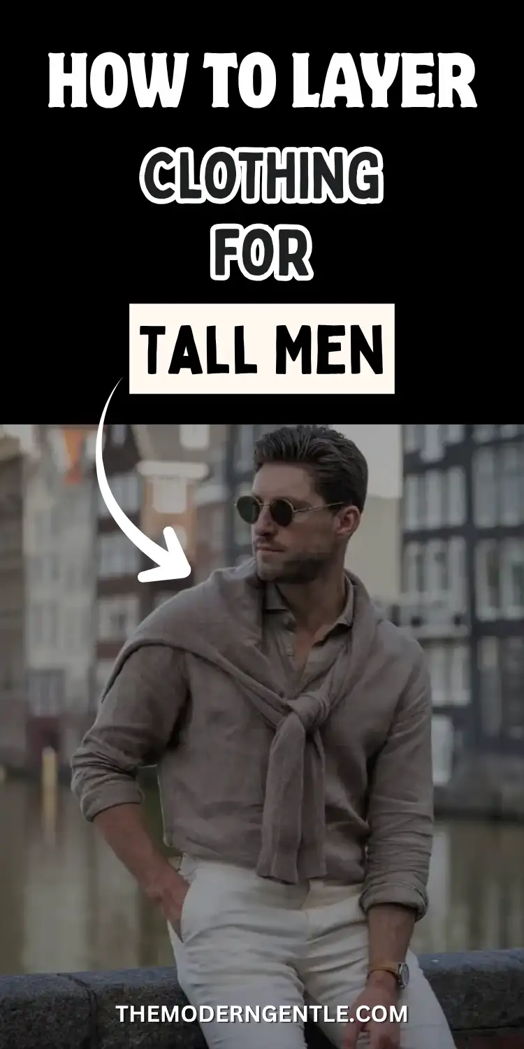 A man in round sunglasses stands beside a European canal in front of tall townhouses, wearing a taupe long sleeve shirt with a matching sweater tied over his shoulders and cream mid to high rise trousers, one hand in his pocket. Bold white text above reads how to layer clothing for tall men, framing this canal side outfit as an example of tall men's fashion and understated proportional style.